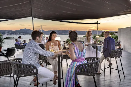 Group of people enjoying an aperitif at CAPOGIRO rooftop, 7Pines Resort Sardinia, with a breathtaking view.
