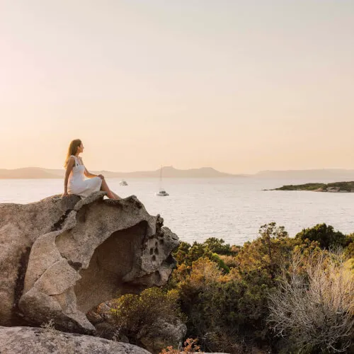 Frau sitzt auf einem Felsen am 7Pines Resort Sardinien und blickt auf das ruhige Mittelmeer