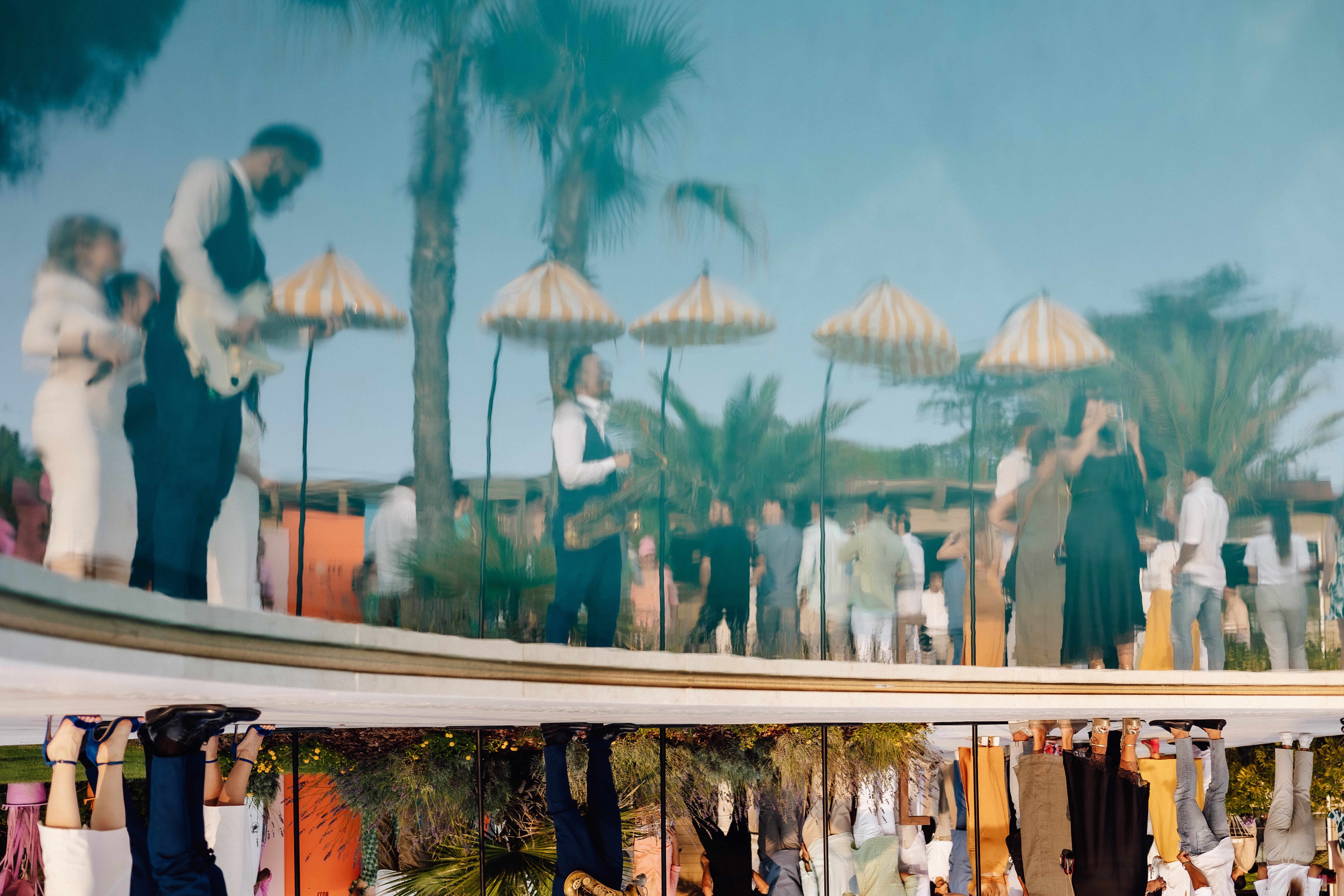 Live musicians and guests at an outdoor event on the terrace of 7Pines Resort Sardinia, with palm trees and striped parasols in the background.