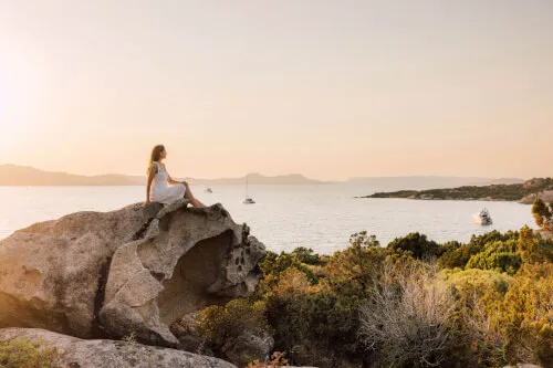 A woman enjoys a sunset view at 7Pines Resort Sardinia, surrounded by sea and rocks.